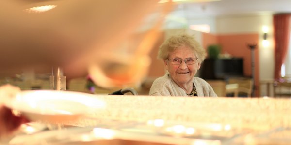Eine Seniorin wartet vor dem Buffet auf ihr Essen und lächelt in die Kamera