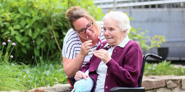 Eine Pflegerin und eine Seniorin riechen gemeinsam an den Blumen im Garten