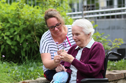 Eine Pflegerin pflückt gemeinsam mit einer Seniorin Blumen im Garten des Seniorencentrums.